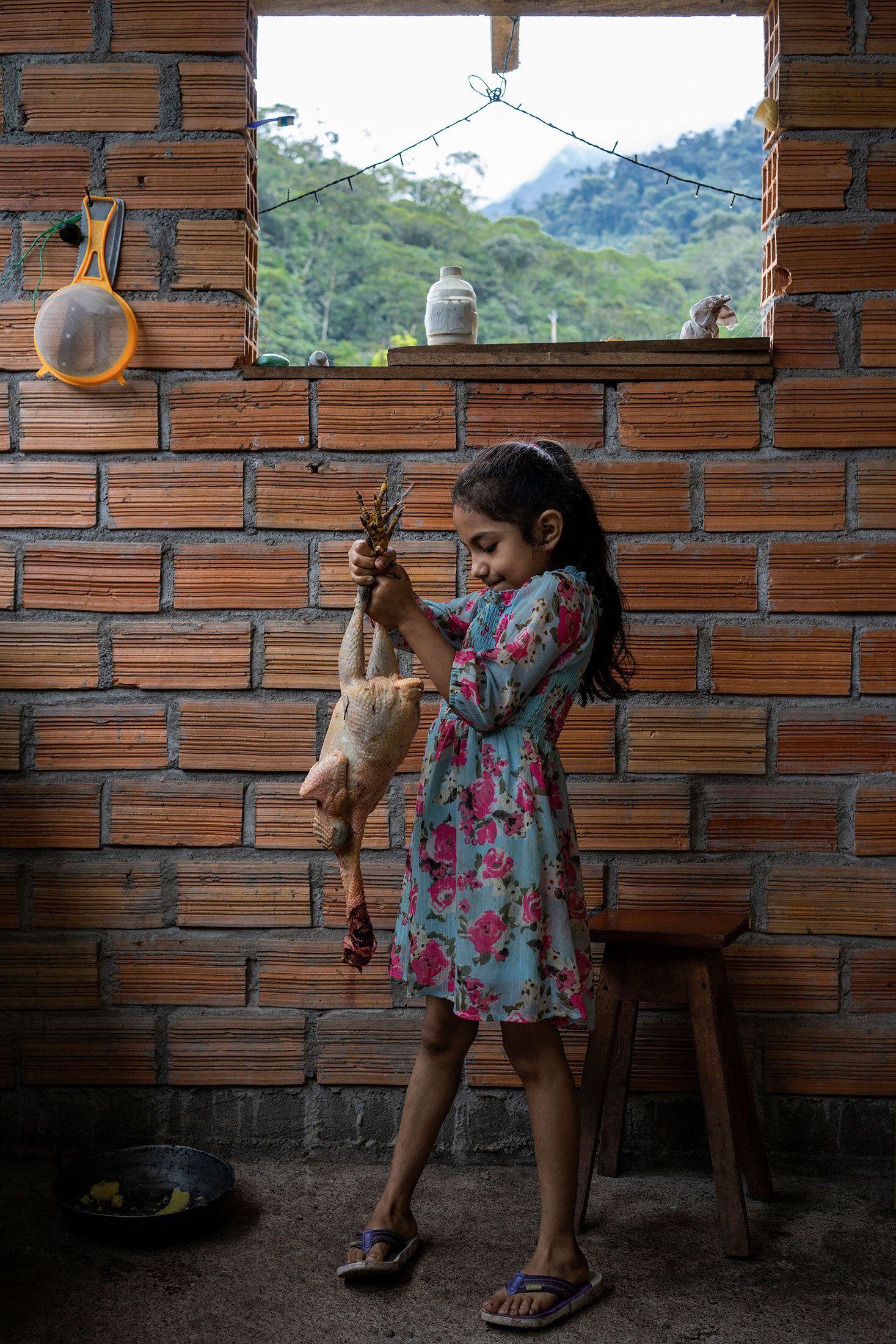A young girl wearing a blue and pink floral dress and flipflops holds a butchered chicken by the legs