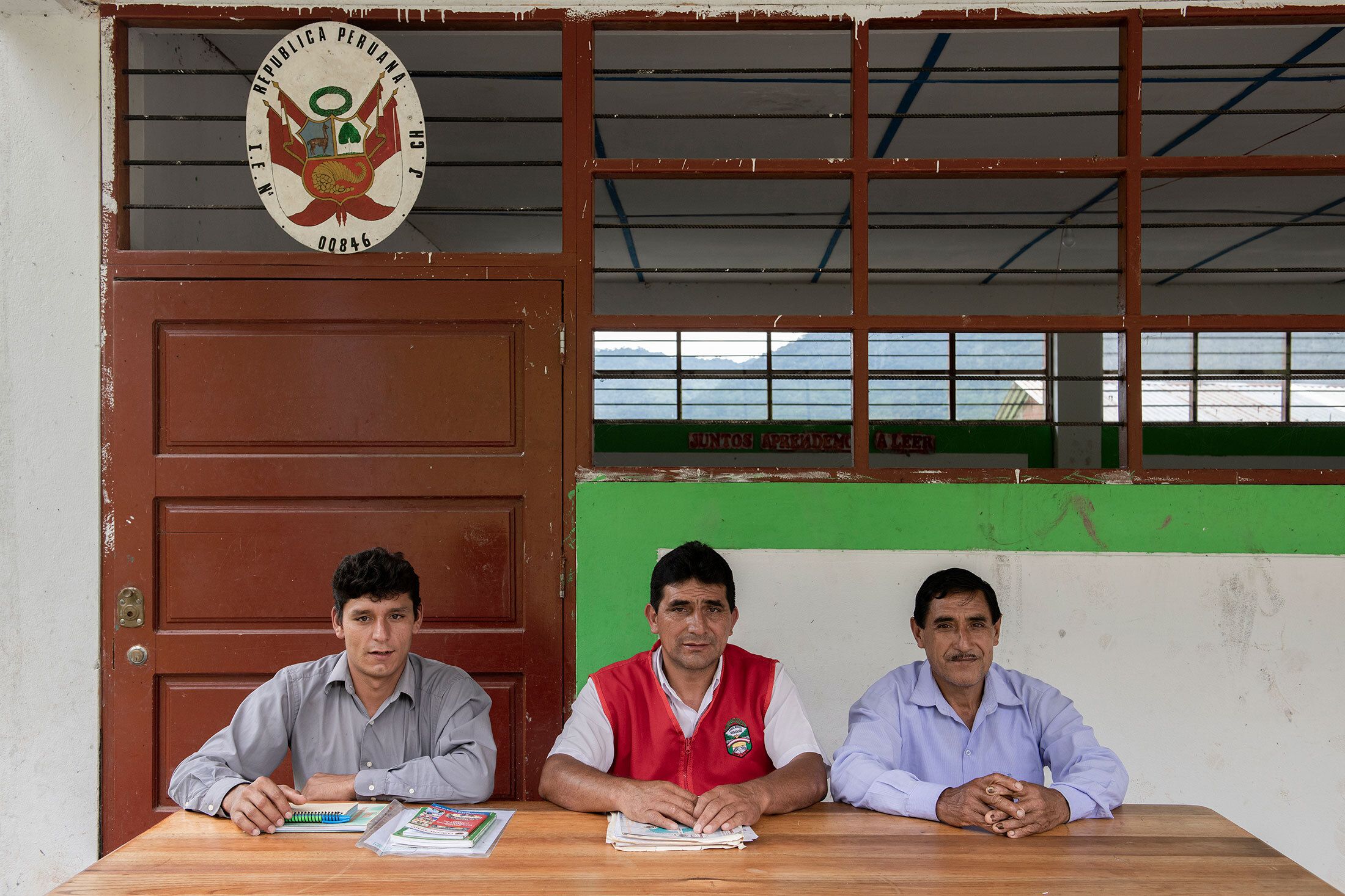 Three men sitting at a table, in front of a weathered building.