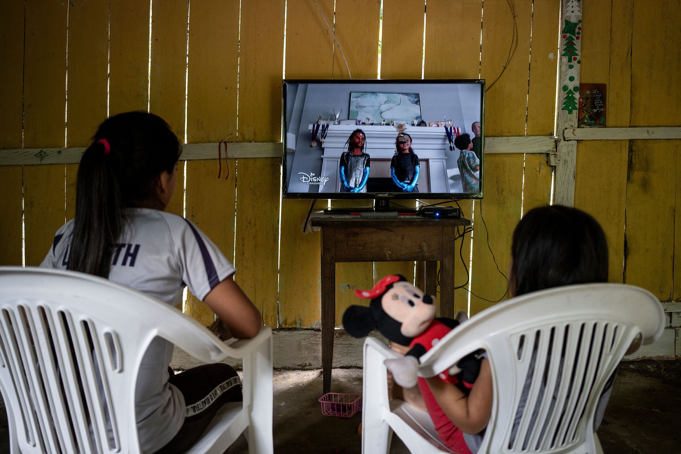Two girls sitting on plastic chairs in front on a TV that plays Disney Channel
