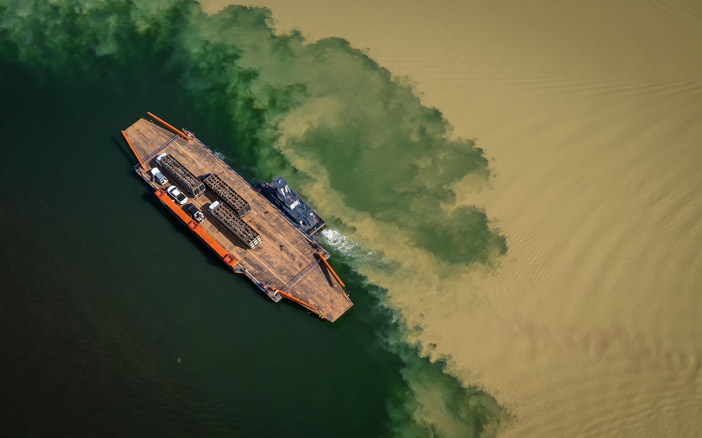 An orange and tan ferry carries three truck-loads of cattle and kicks up silt in the emerald-green river.