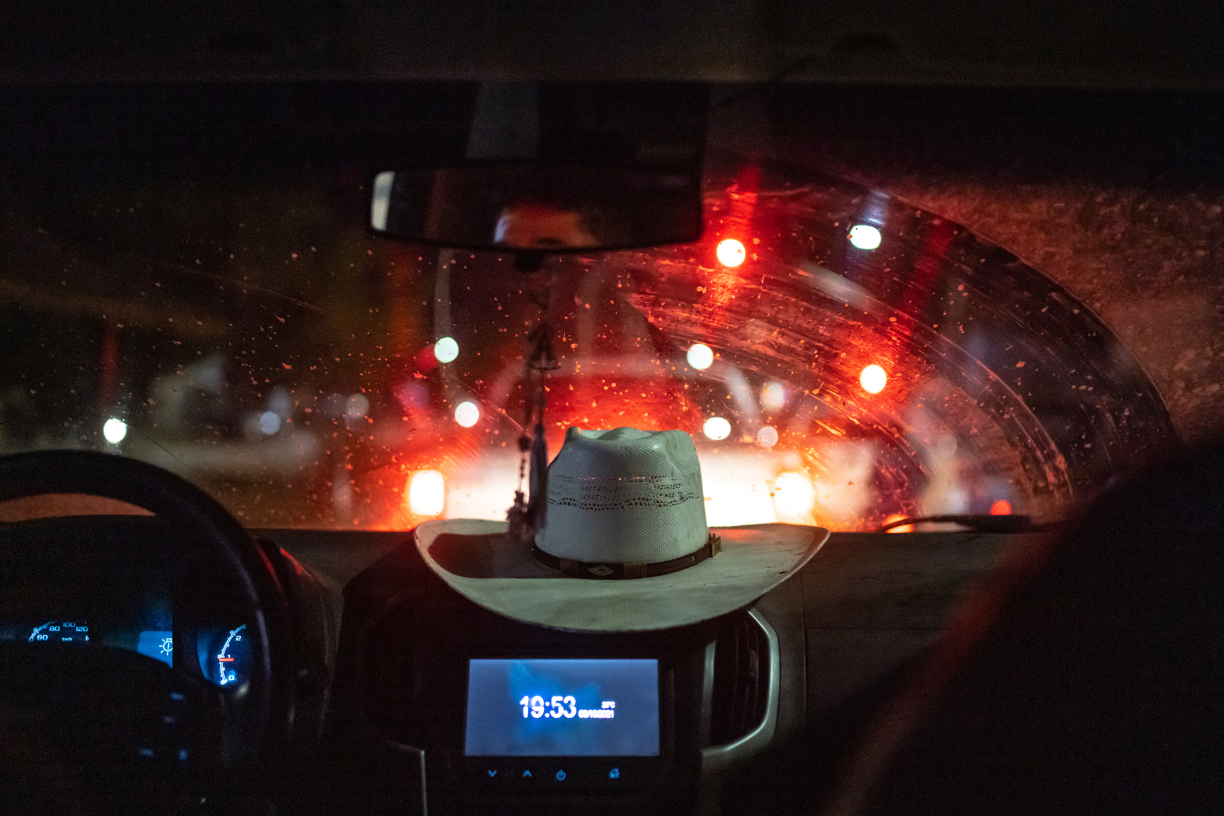 A white cowboy hat sits on the dashboard of a car at night. The red tail lights of the vehicle ahead illuminate an otherwise dark car interior.