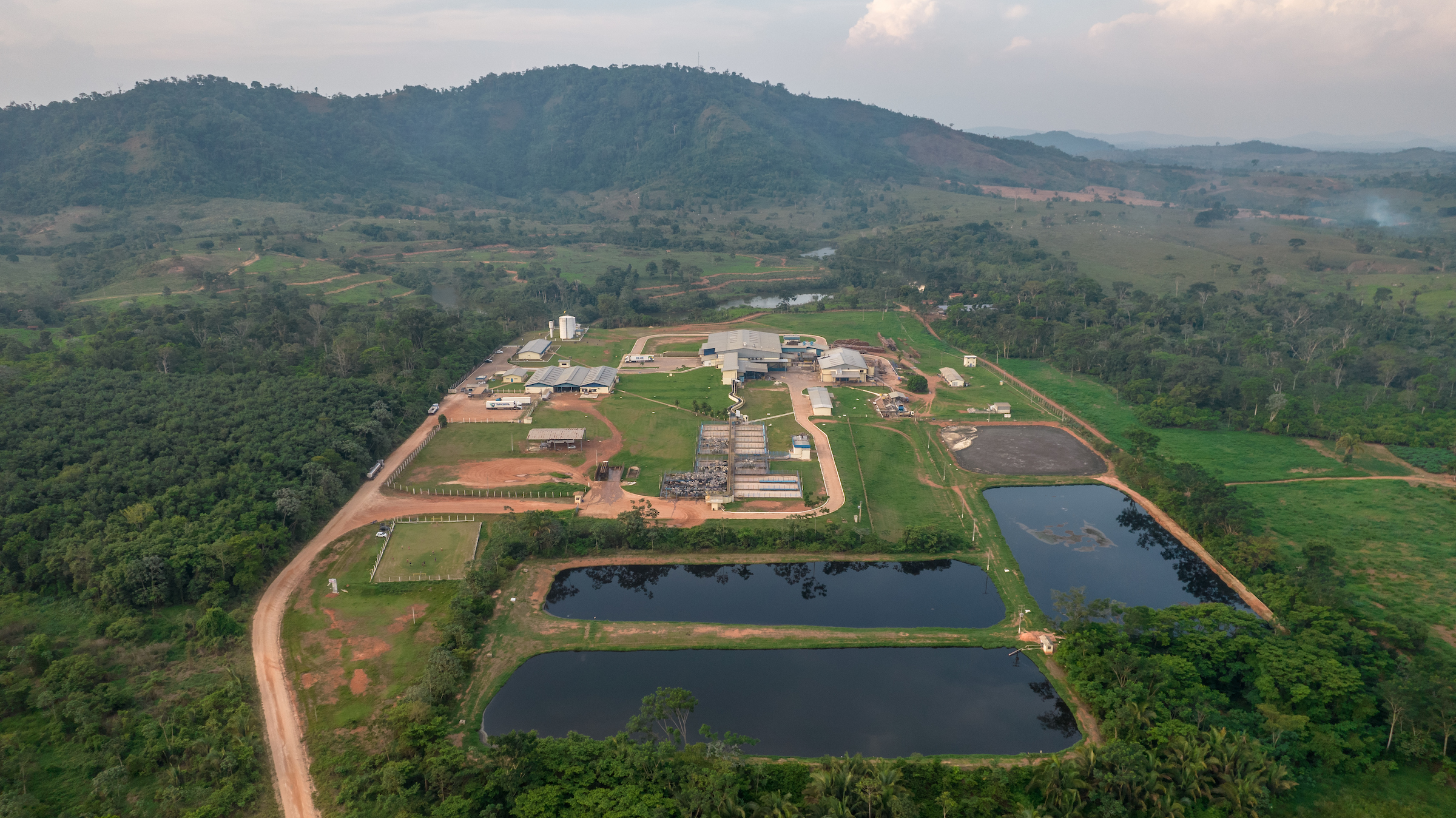 An aerial view of a compound with cattle pens, warehouses, and reservoirs. A fire smoulders in the distance.