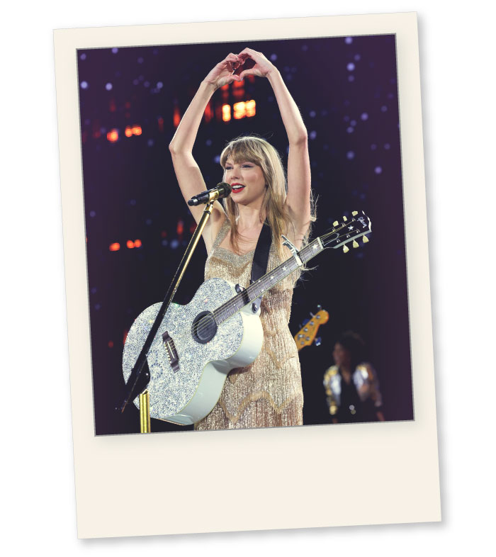 Taylor Swift performing during one of the Eras Tour shows, in East Rutherford, New Jersey. Swift is wearing a golden dress and has a silver, glittery guitar. She&rsquo;s in front of a microphone and her arms are over her head, she makes a heart shape with her hands while smiling. The background is blurry but you can see small lights. The picture is cropped as a polaroid and has a blue/purple color to it.