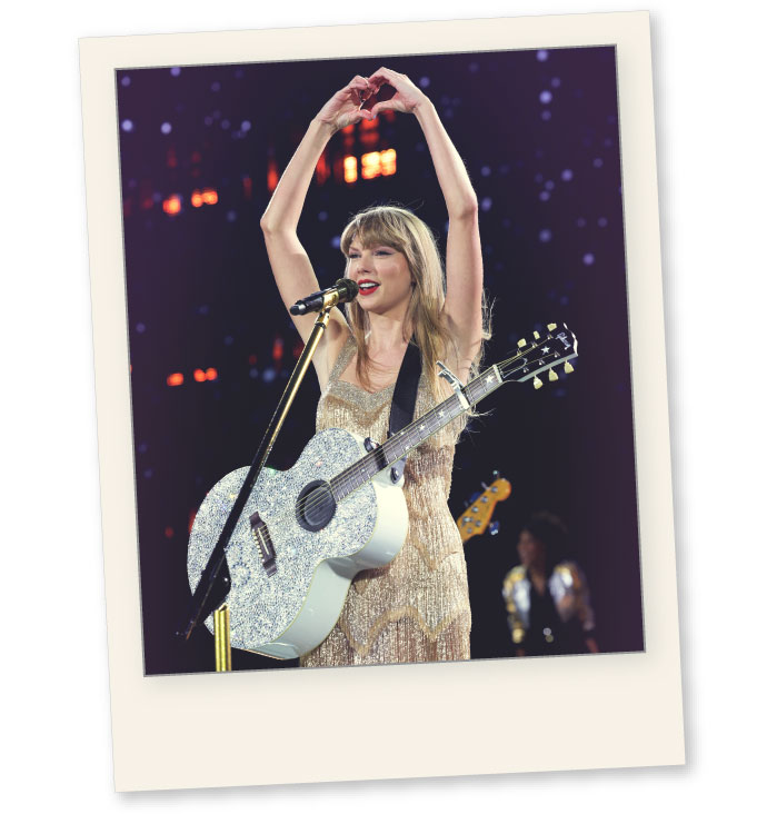 Taylor Swift performing during one of the Eras Tour shows, in East Rutherford, New Jersey. Swift is wearing a golden dress and has a silver, glittery guitar. She&rsquo;s in front of a microphone and her arms are over her head, she makes a heart shape with her hands while smiling. The background is blurry but you can see small lights. The picture is cropped as a polaroid and has a blue/purple color to it.