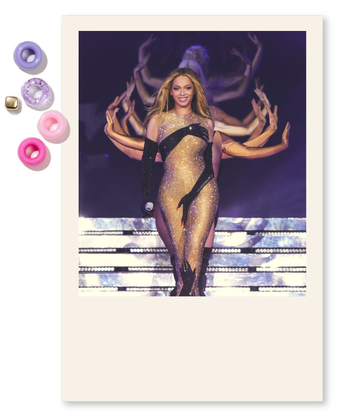 A picture of Beyonce performing during her renaissance tour. She is smiling and her hair looks flowy. Behind her are dancers, waving their hands on each side of Beyonce as she walks. Beyonce is wearing a shiny jumpsuit with hands depicted on it, and black gloves. The picture is cropped as a polaroid.