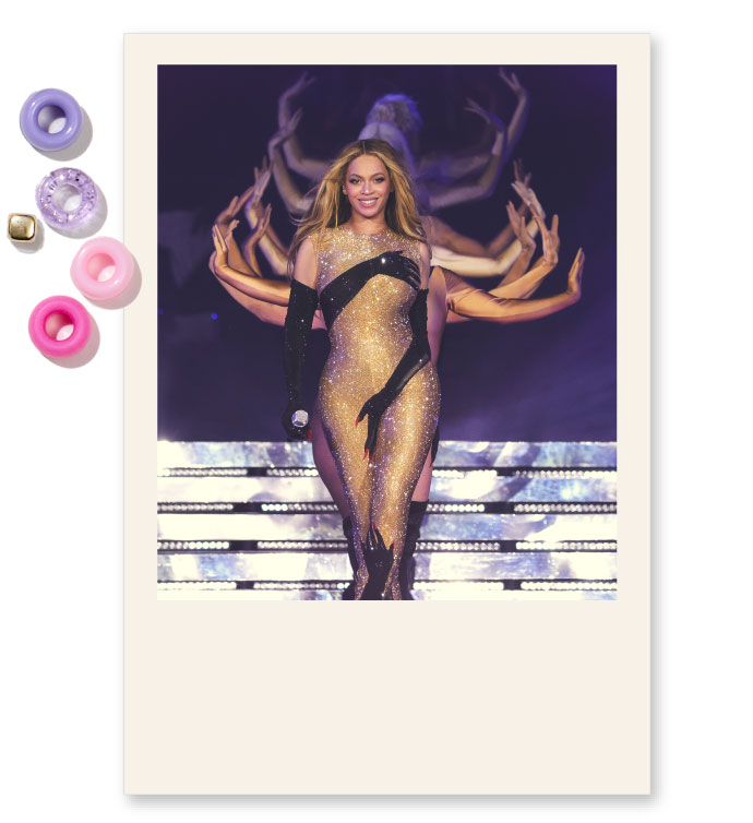 A picture of Beyonce performing during her renaissance tour. She is smiling and her hair looks flowy. Behind her are dancers, waving their hands on each side of Beyonce as she walks. Beyonce is wearing a shiny jumpsuit with hands depicted on it, and black gloves. The picture is cropped as a polaroid.