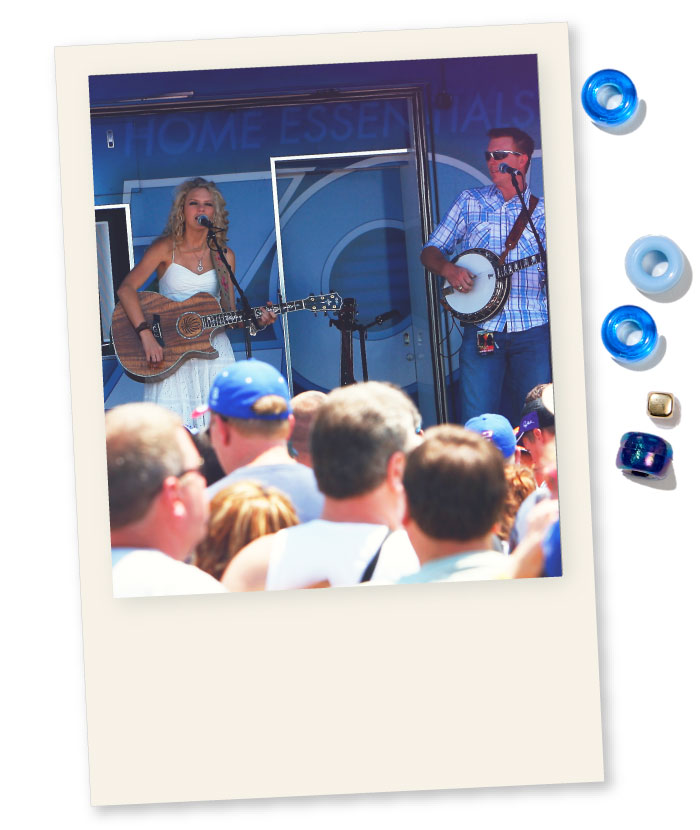 a Young Taylor Swift playing the guitar and singing on a stage where the backdrop reads &ldquo;home essentials&rdquo;. She is accompanied by a man wearing sunglasses who&rsquo;s playing the banjo. The foreground of the picture features multiple people listening to the performance.