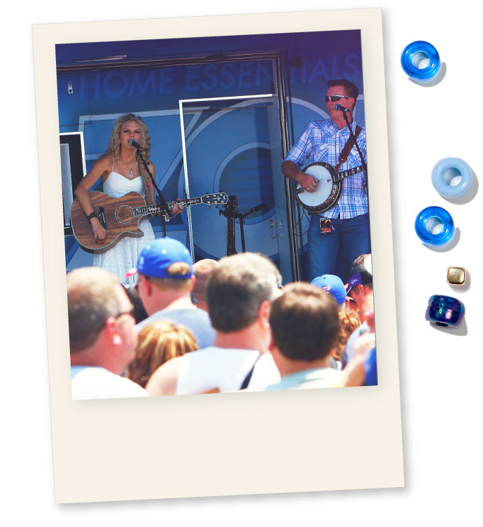 a Young Taylor Swift playing the guitar and singing on a stage where the backdrop reads &ldquo;home essentials&rdquo;. She is accompanied by a man wearing sunglasses who&rsquo;s playing the banjo. The foreground of the picture features multiple people listening to the performance.